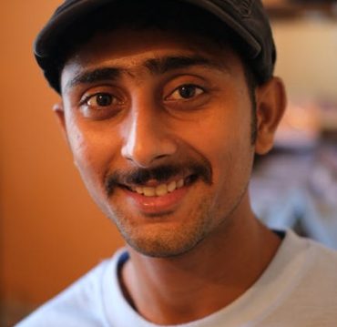 Close-up portrait of a young man wearing a cap, smiling warmly indoors.