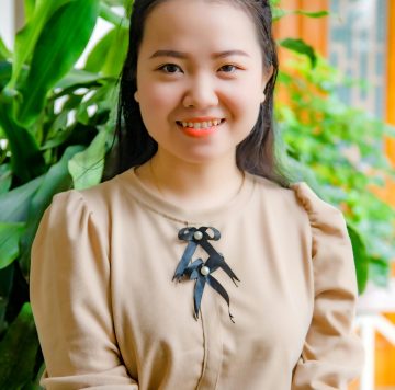 A smiling woman wearing a beige top, sitting in front of green plants. Bright and cheerful.